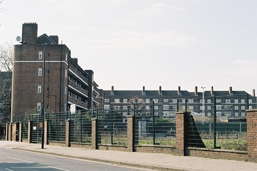 A view of the Robin Hood Housing Estate, London. It is built out of dark brown brick. In the foreground by the pavement is a fence bordering the grassy recreation are. The face is made up of a short wall, brick posts and barbed wire that goes across the image. The sky is grey and the trees have no leaves.